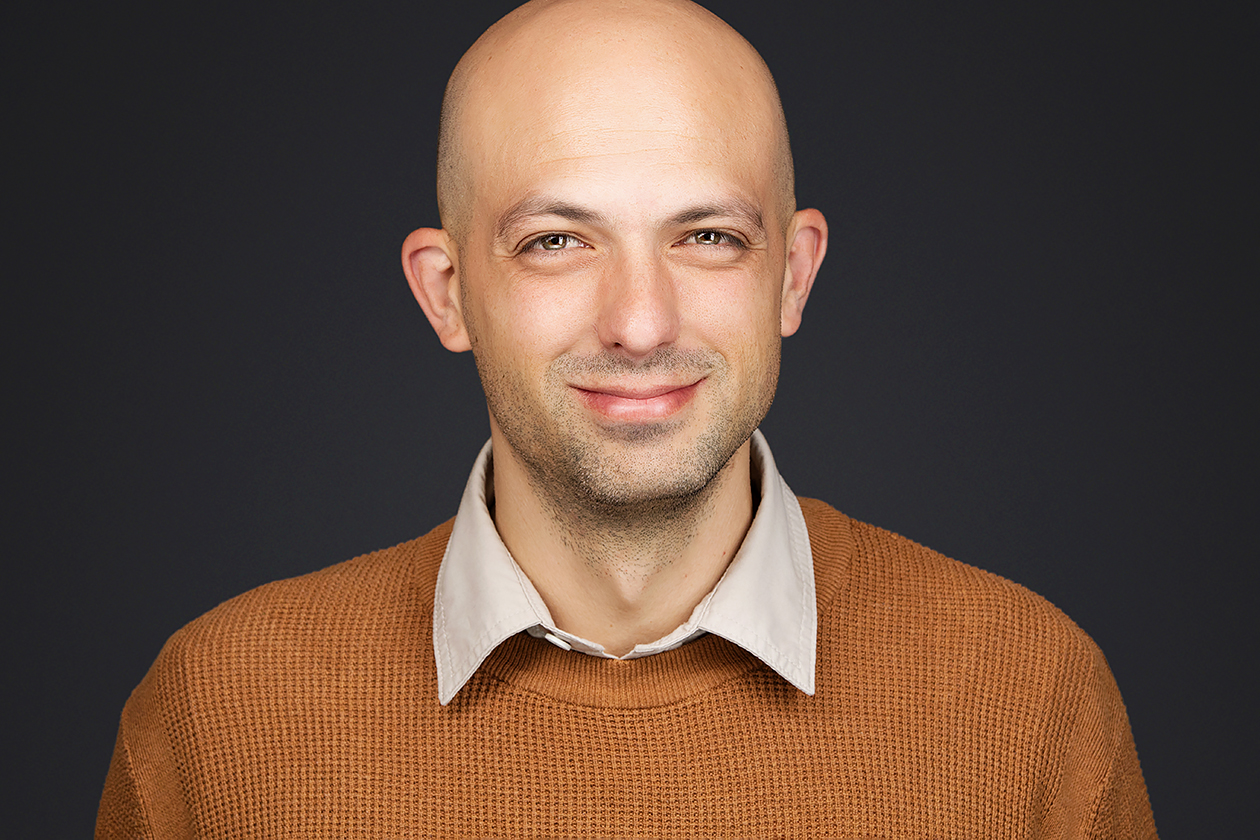 Executive headshot of a middle-aged white man in a mustard pullover, smiling confidently